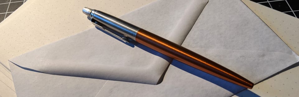 A pen (a Parker Jotter, orange-ish accent), laying on its side on top of some stationary.